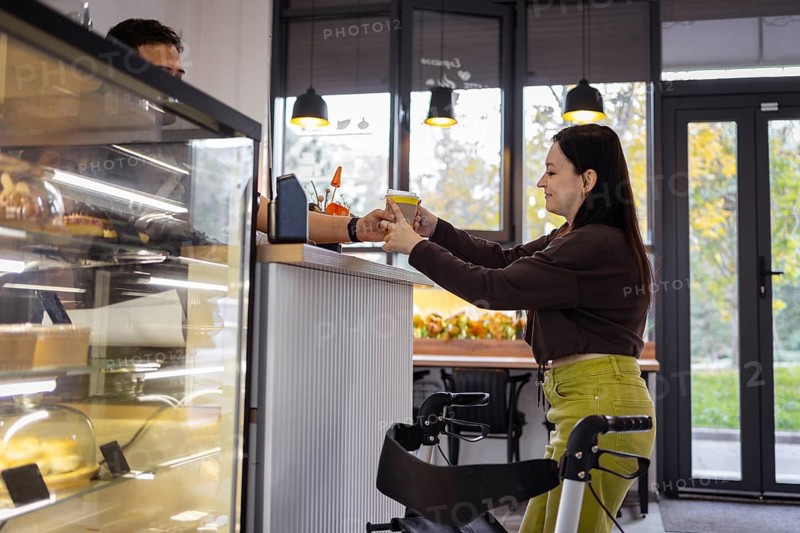 A woman with cerebral palsy receives a coffee cup from a barista at a cafe counter. A display case with pastries is visible, and the warm ambiance is enhanced by pendant lights and autumn decor