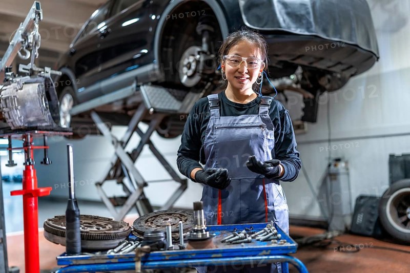 Confident young female mechanic wearing gloves and protective glasses, working on car parts in a professional auto repair shop