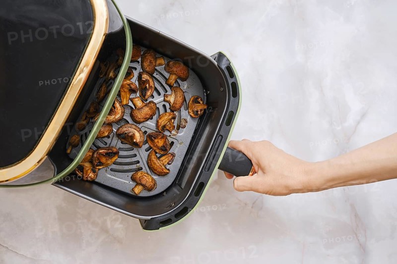 A hand holds the basket of an air fryer that contains cooked champignon mushrooms. The mushrooms are brown and appear crispy. The kitchen counter has a neutral surface, indicating a home cooking scene