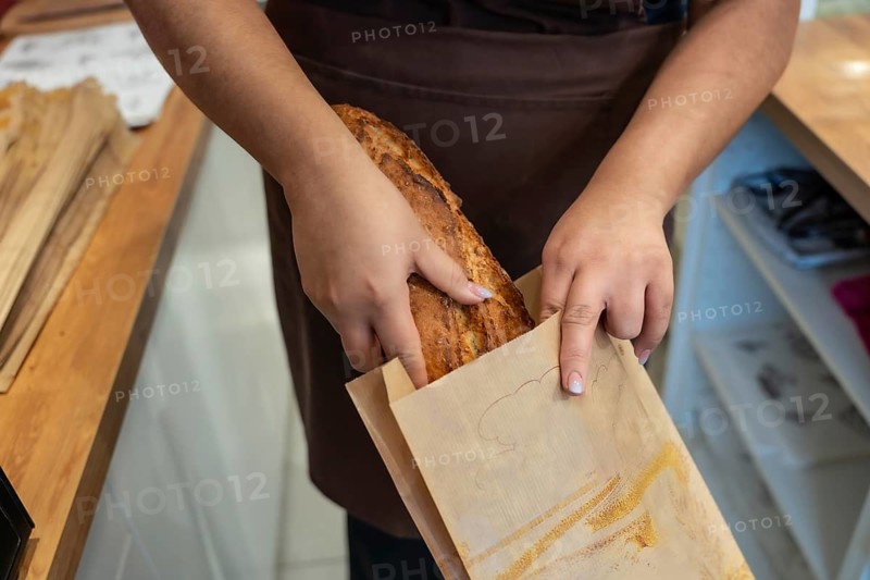 Baker's hands carefully placing a fresh, crusty artisan bread loaf into a brown paper bag, emphasizing traditional craftsmanship and sustainable packaging in a bakery setting