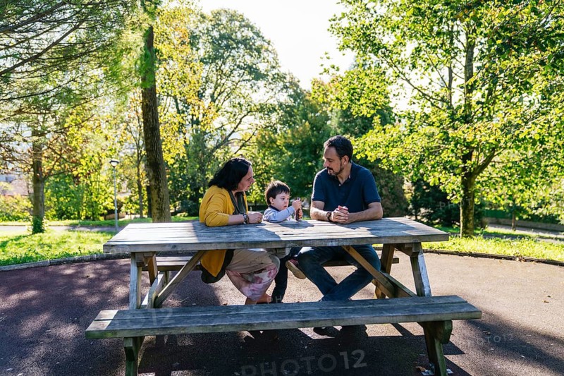 A family of three enjoys quality time at a park picnic table. Surrounded by lush greenery, the parents and child with Down syndrome share a moment of connection and relaxation outdoors