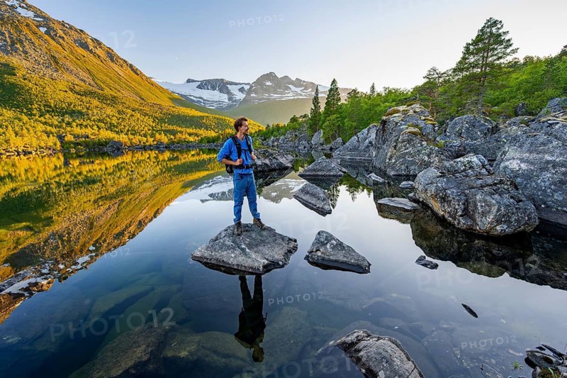 Young man standing on a rock in water, enjoying the landscape, picturesque mountain scenery in the Innerdalen high valley, mountain peaks reflected in Lake Innerdalsvatna, in the evening light at sunset, Trollheimen Mountain Area, Sunndal, Møre og Romsdal, Norway