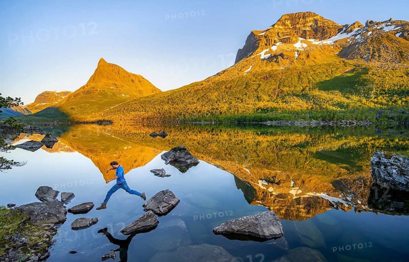 Young man jumping from stone to stone in water, picturesque mountain landscape in the Innerdalen high valley with Innerdalstårnet summit, mountain peaks reflected in Lake Innerdalsvatna, in the evening light at sunset, Trollheimen Mountain Area, Sunndal, Møre og Romsdal, Norway