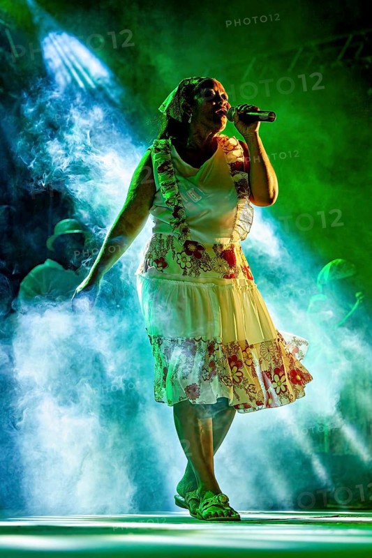 Woman singing on stage surrounded by mystical green light and fog in floral print dress, a band plays and dances at a festival in Petrolina in Brazil