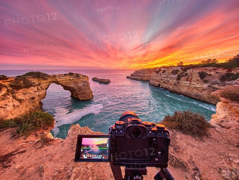 Dramatic sunset at Arco de Albandeira in Algarve, Portugal, with a camera on tripod above the limestone sea arch, pink sky, and Atlantic water