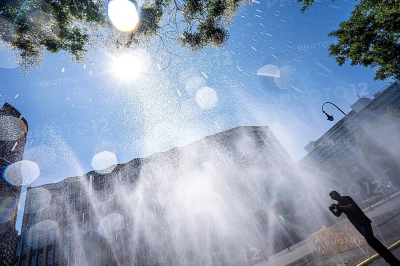 Cooling Cologne, campaign by the city of Cologne to cool down places exposed to heat, here Rudolfplatz in the city center, at just under 37°C during the day, a spray hose splashes fine water droplets that cool down the immediate area a few degrees, an action carried out when there is extreme heat stress, to adapt the urban climate to climate change, passers-by enjoy the cooling moment, plus tips on how to deal with extreme Hitze, North Rhine-Westphalia, Germany