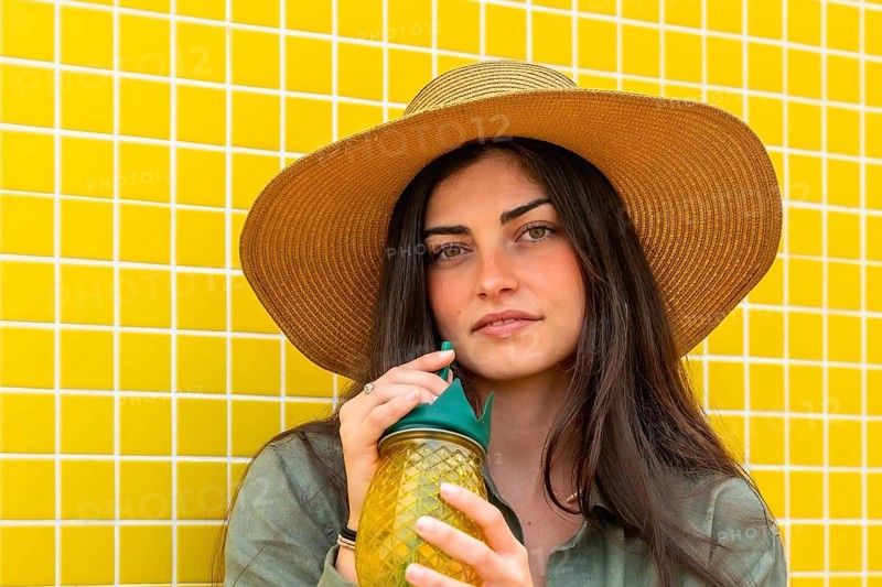 Woman wearing a straw hat and a casual green shirt. Drinking a cold beverage from a pineapple shaped glass. Standing in front of a vibrant yellow tiled wall. Enjoying the summer and vacation lifestyle