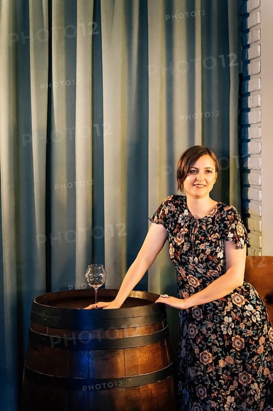 A smiling woman in a dark floral dress stands in a bar interior next to a wooden wine barrel with a glass placed on it. Warm lighting, tall curtains, and rustic decor create a cozy hospitality atmosphere suitable for wine tasting, bar service, or restaurant promotion. Blonde with a glass in the wine bar near the wine barrel