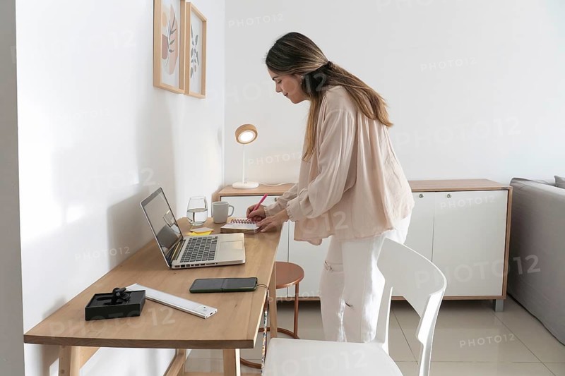 Woman leaning over wooden desk writing notes in spiral planner with pink pen, laptop open beside her in bright minimalist home office