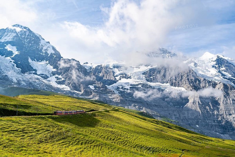 Jungfrau Railway to Jungfraujoch runs through green meadows, above the Kleine Scheidegg stop, rack railway, in the back mountains with peaks Eiger, Mönch and Jungfrau, Jungfrau region, Bernese Oberland, Switzerland