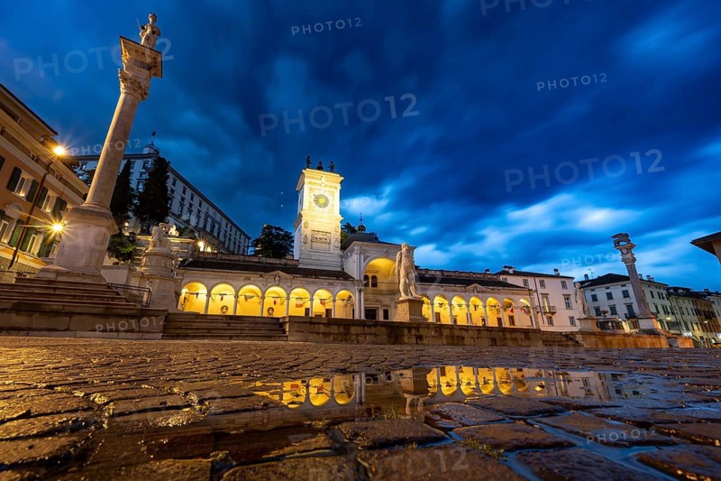 Illuminated main square Piazza della Libertà with Loggia e Tempietto di San Giovanni and clock tower, Castello di Udine behind, blue hour, reflection in water puddle, dramatic sky, old town, Udine, Friuli-Venezia Giulia, Italy