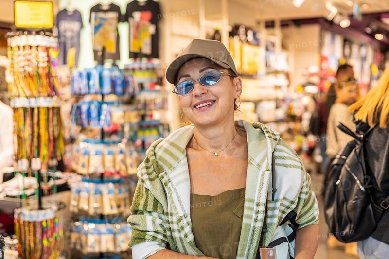 Middle-aged Caucasian woman with glasses and a cap stands in a souvenir shop. Colorful merchandise and displays are visible in the background