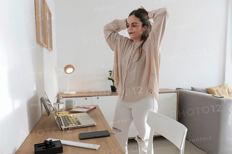 Woman taking a break from work, stretching arms and tying her hair up, standing beside desk with open laptop in bright minimalist apartment