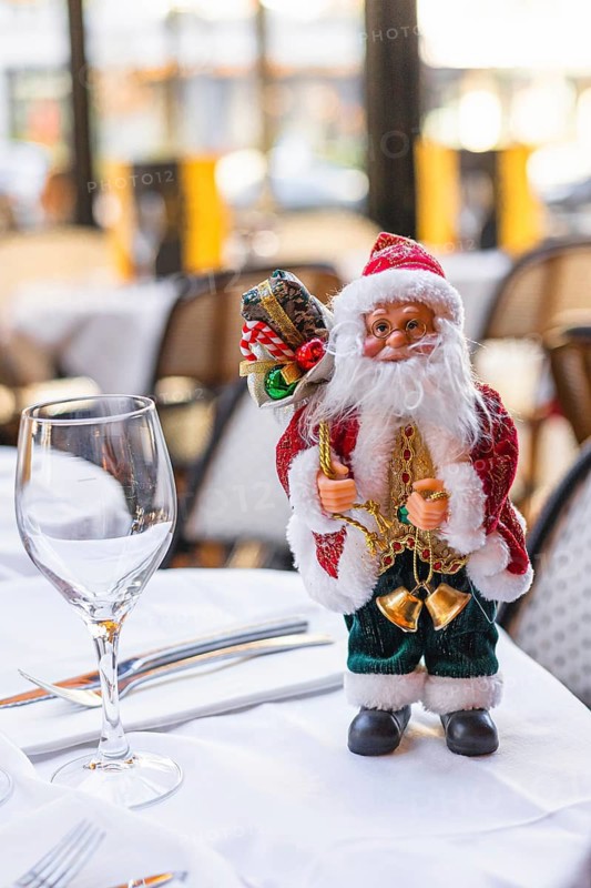 A decorative Santa Claus figurine stands on a table. The Santa has a red suit, white beard, and holds gifts. A wine glass is placed beside him