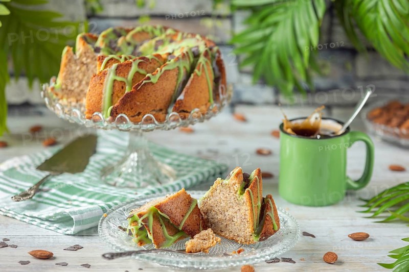 A sliced cake and pieces of cake with green icing next to a cup of coffee and almonds