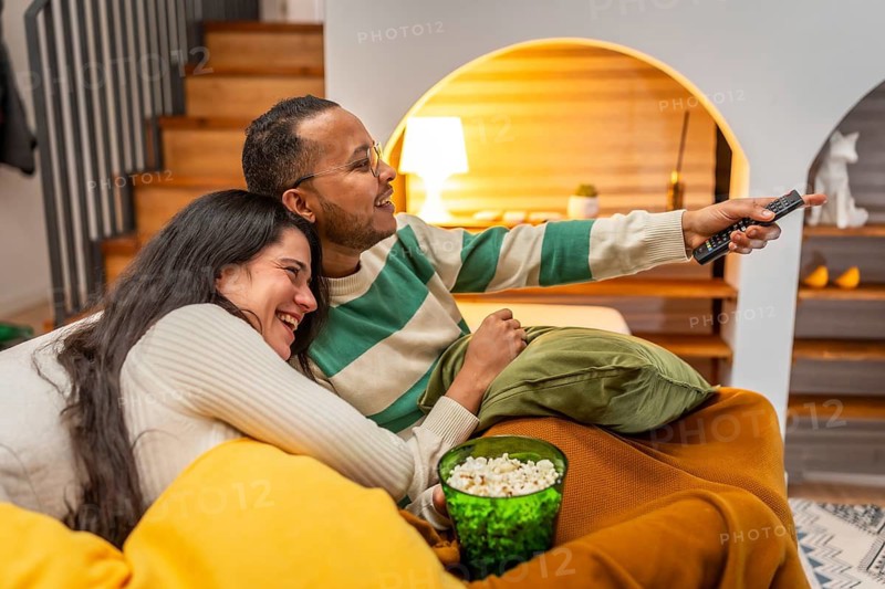 Happy smiling multiracial couple cuddled on a cozy sofa, sharing popcorn and a remote while watching a movie or show in their relaxed, intimate living room at home