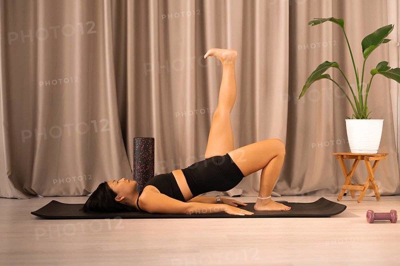 Woman stretching on a yoga mat indoors, next to a potted plant and curtain