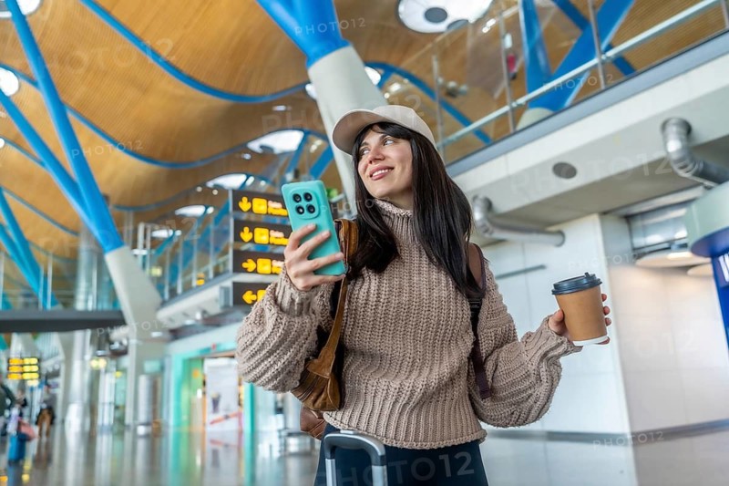 Young woman carrying luggage, holding a mobile phone and coffee cup, smiling and looking away while navigating through a modern airport terminal during her solo travel journey