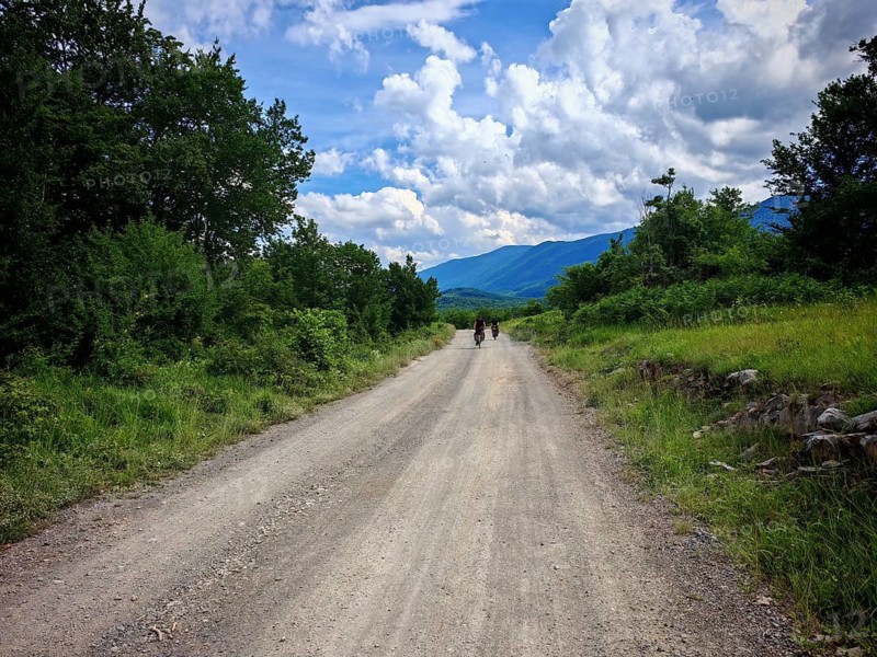 Two cyclists ride on a dusty track, path lined with lush vegetation, mountains in the background, under a cloudy sky, hinterland Croatia, near Kvarner Gulf Bay, Croatia