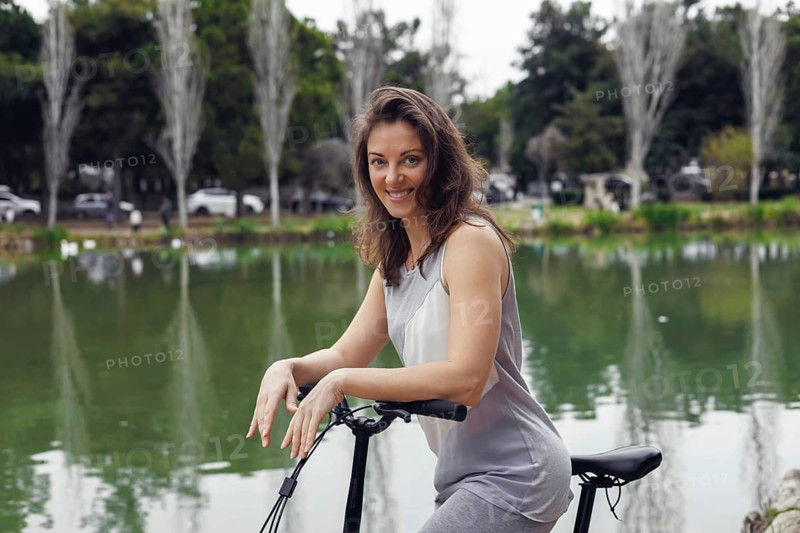 A young Caucasian woman with long brown hair smiles while riding an electric scooter by a serene lake surrounded by trees