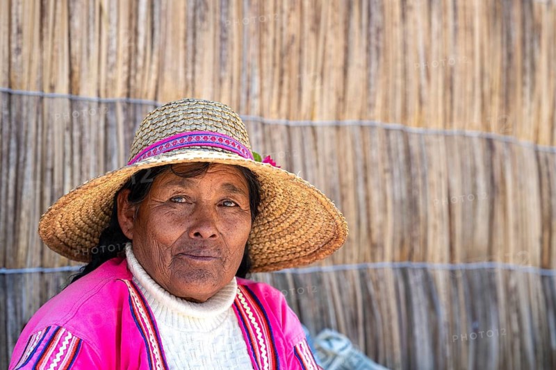 A friendly elderly woman wearing a hat, a member of the indigenous population, a descendant of the Uros (Uru), on a reed island in front of a house wall made of totora reeds on Lake Titicaca, the Altiplano, southern Peru, Peru, South America