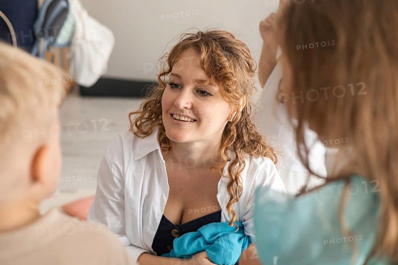 A young Caucasian woman with curly brown hair interacts with children in a cozy home setting during a live music event. The atmosphere is warm and inviting