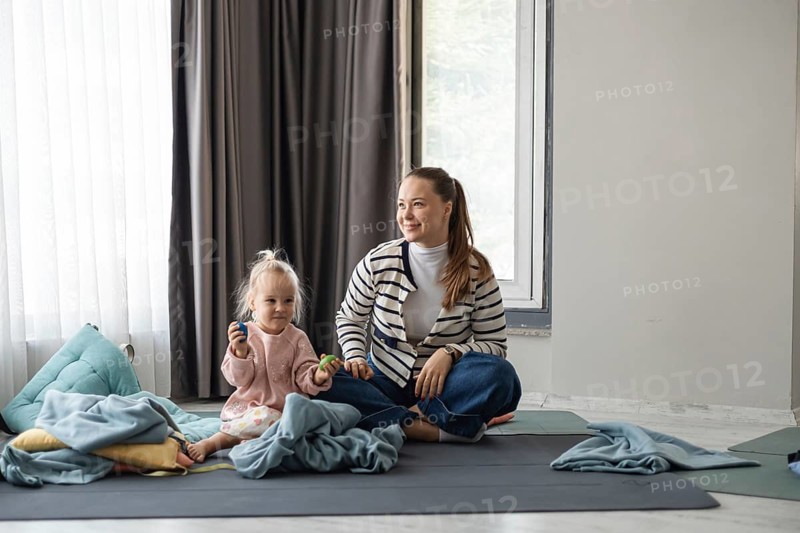A young Caucasian woman with long brown hair sits on a mat with a toddler girl. The girl has blonde hair and is playing with toys. They are surrounded by blankets in a cozy home setting