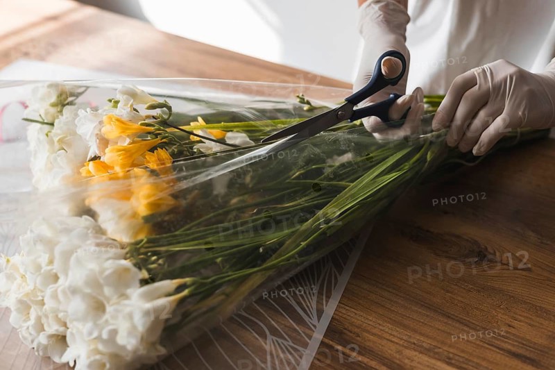 A person in gloves is cutting green herbs with scissors on a wooden surface. Colorful flowers are nearby, enhancing the cheerful atmosphere of a bright flower shop setting