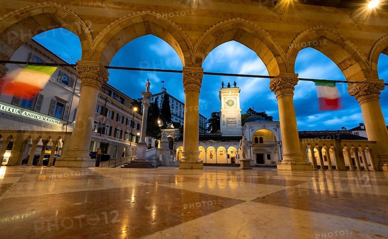 Arcades of the Loggia del Lionello, illuminated main square Piazza della Libertà with Loggia e Tempietto di San Giovanni and clock tower, Castello di Udine behind, blue hour, dramatic sky, old town, Udine, Friuli-Venezia Giulia, Italy