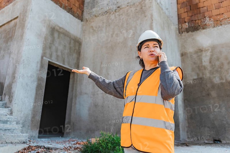 A young Hispanic woman in a safety vest and hard hat gestures while speaking on a phone at a construction site. The background shows unfinished concrete walls