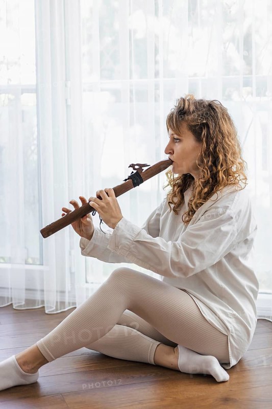 A woman in a white shirt playing a wooden flute while seated on a wooden floor