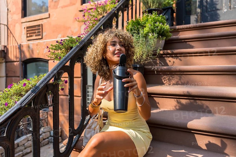 Stylish young woman with curly blonde hair staying hydrated on a residential brownstone stoop in new york city during a warm summer day