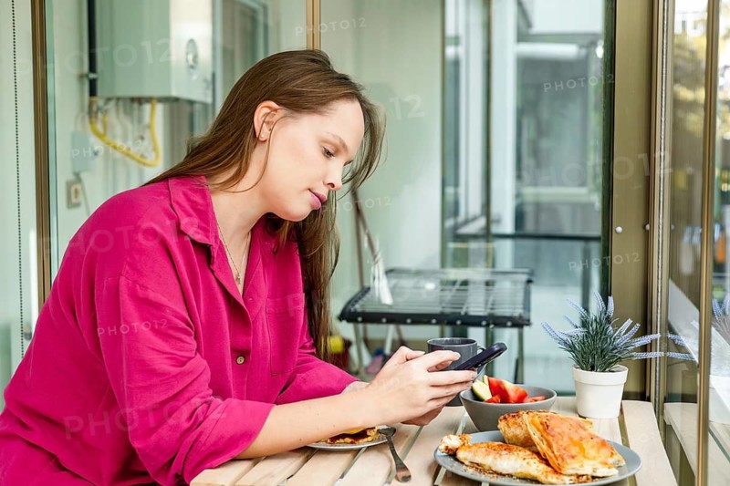 A young Caucasian woman with long brown hair sits at a table, enjoying a meal. She wears a pink robe and appears relaxed in a bright, modern setting