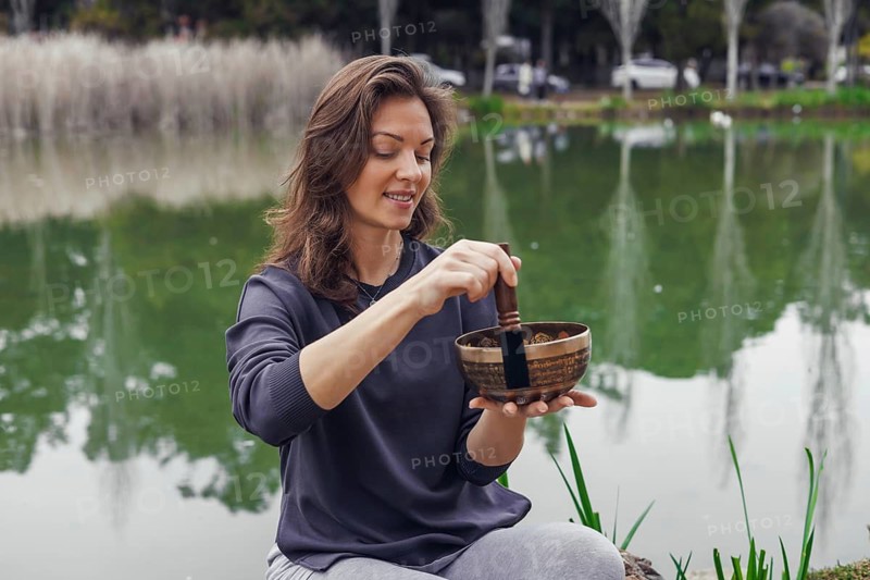 A young Caucasian woman with brown hair sits by a serene lake, holding a wooden bowl filled with colorful ingredients. Green reeds surround the water