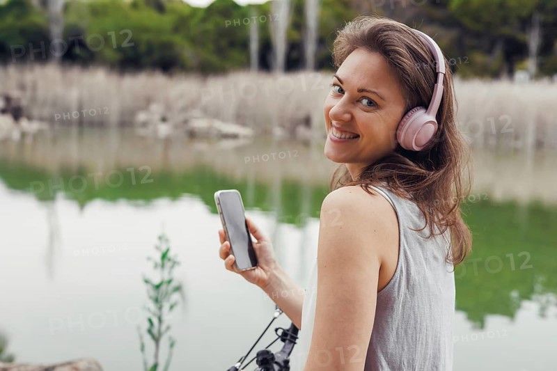 A young Caucasian woman with brown hair wearing headphones stands by a lake, checking her smartphone next to a bicycle