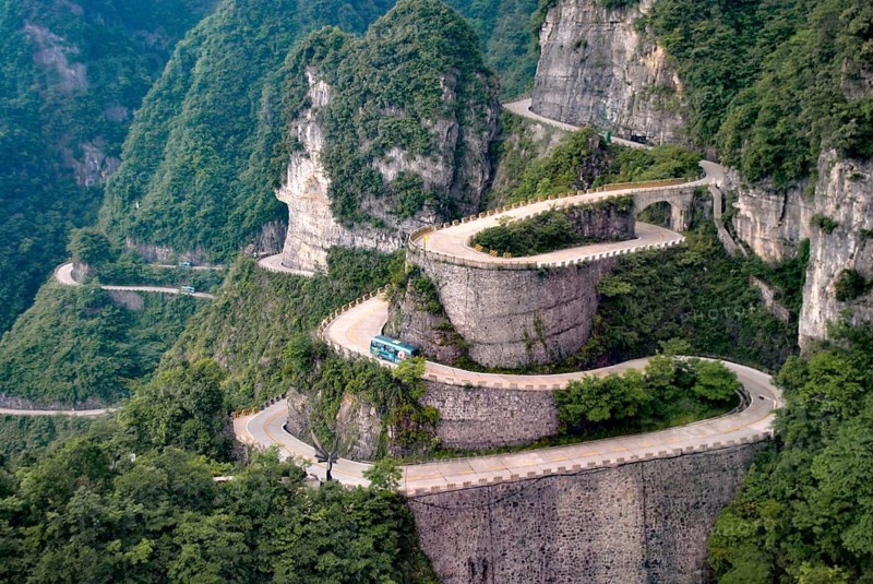 Heaven Linking Avenue on Tianmen Mountain, China, A winding road with sharp turns amidst lush green mountains and stone walls, showcasing a scenic and dramatic landscape