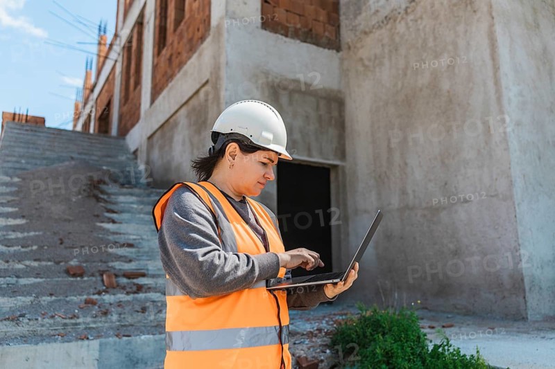 Woman in a hard hat and safety vest uses a laptop on a construction site. The background features unfinished buildings and stairs