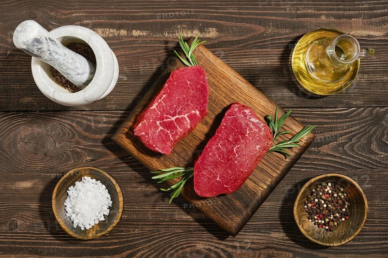 Two pieces of raw beef steak on wooden board surrounded by fresh herbs. There are bowls of salt and pepper nearby. Olive oil is present along with stone mortar