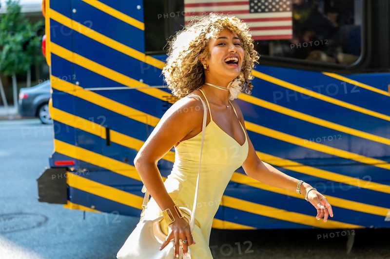 Cheerful young woman with curly blonde hair walking on a sunny day in manhattan, expressing joy and confidence while experiencing the vibrant city life near a public transit bus