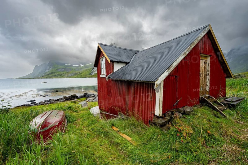 Red wooden houses in green mountain landscape with dramatic clouds, Vindstad, Reinefjord, Lofoten, Norway
