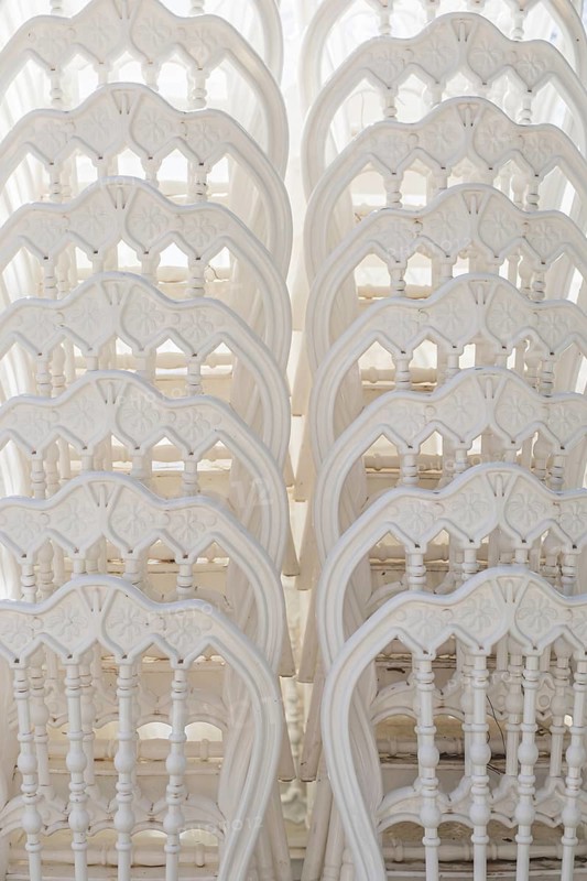 A stack of white wooden chairs arranged closely together. The chairs have intricate designs and a smooth finish, creating a clean and elegant background texture
