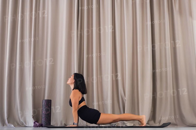 Woman practicing yoga stretching pose on a mat with neutral curtains in the background