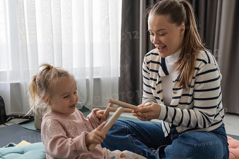 A young Caucasian woman and a little girl play with wooden music sticks at home. They are smiling and enjoying a sensory music activity together