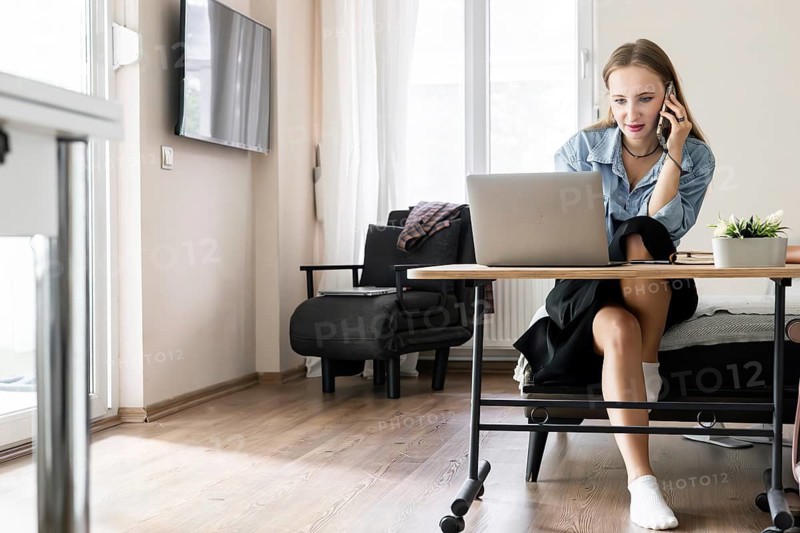 A young Caucasian woman with long brown hair sits at a desk, using a laptop and talking on a phone in a modern living room
