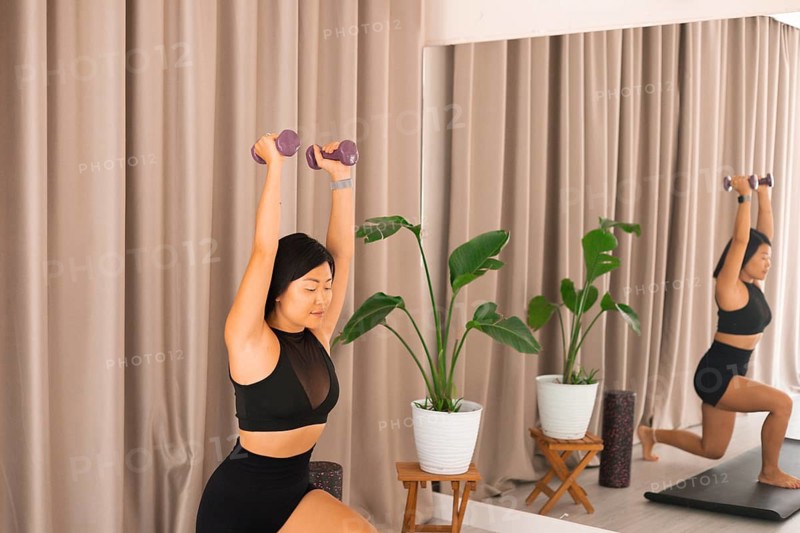A woman lifts dumbbells in a focused position with a mirrored reflection and a serene background