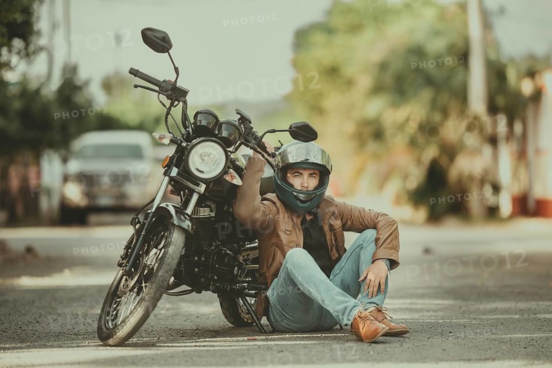 Portrait of handsome motorcyclist sitting next to his motorcycle on the road. Portrait of young motorcyclist leaning against his motorcycle on the road