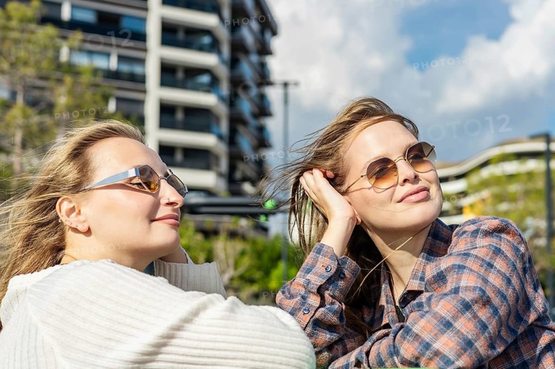 Two young women enjoying a sunny day outdoors. One has long blond hair and wears sunglasses. The other has long brown hair and a plaid shirt. Modern buildings in the background