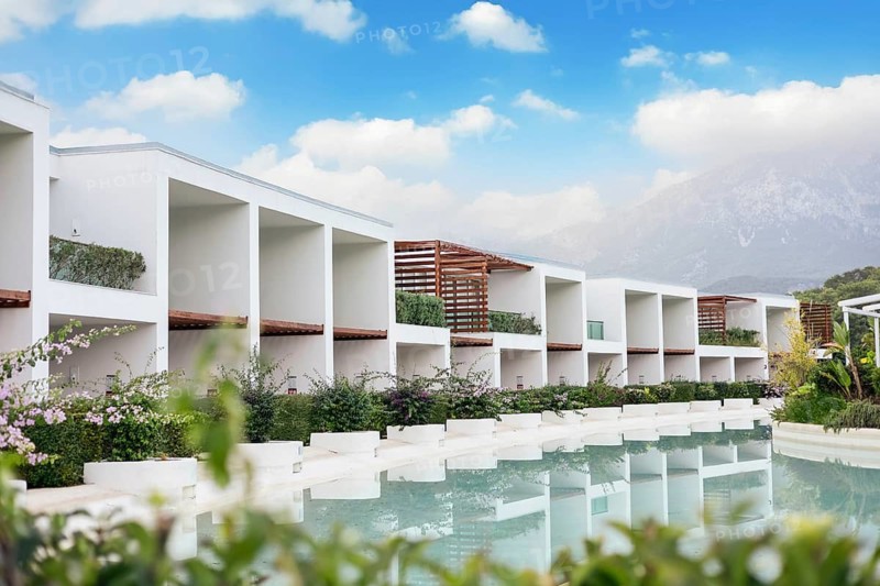 Modern white resort buildings beside a tranquil pool, surrounded by greenery and under a blue sky with clouds. Antalya, Turkiye