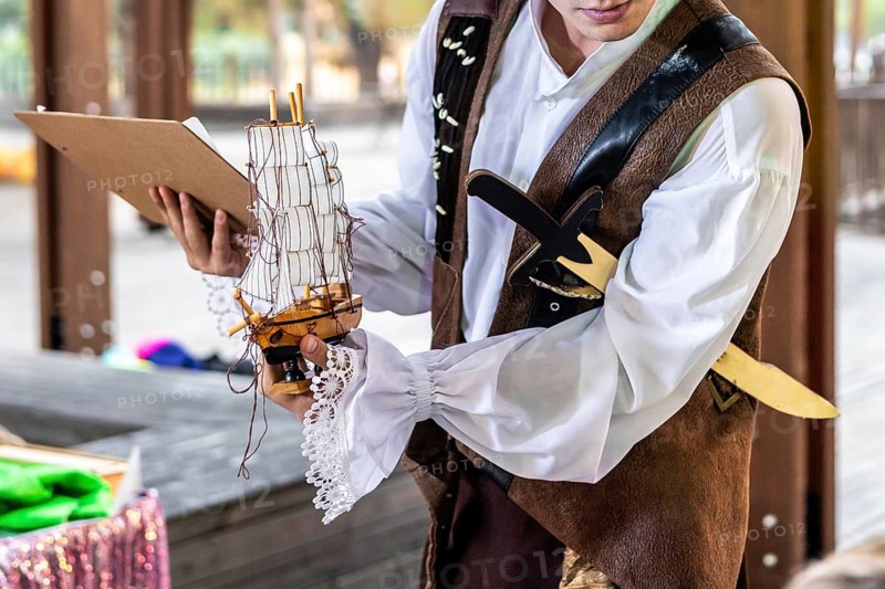 A young Caucasian man in a pirate costume engages a group of children during a storytelling session. The children are attentive and excited