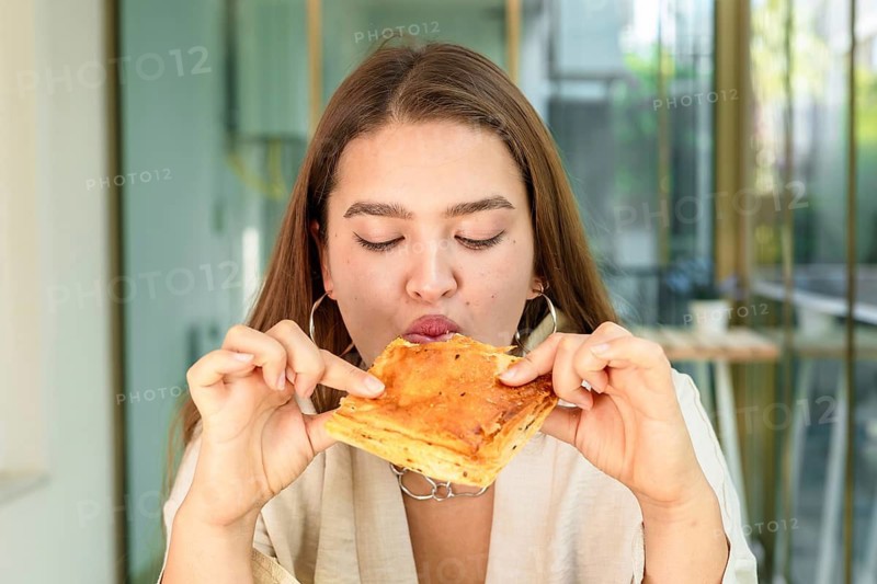 Young Caucasian woman with long brown hair enjoys a pastry in a bright, modern cafe. She is focused on the food, creating a casual dining atmosphere